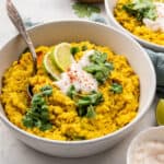 A bowl of kitchari topped with yogurt, cilantro, and limes, with a spoon in it, with a bowl of kitchari and a bowl of cilantro in the background