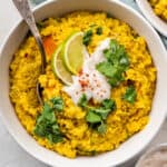 Close up overhead of a bowl of kitchari topped with cilantro, yogurt, and lime wedges, with a spoon in it.