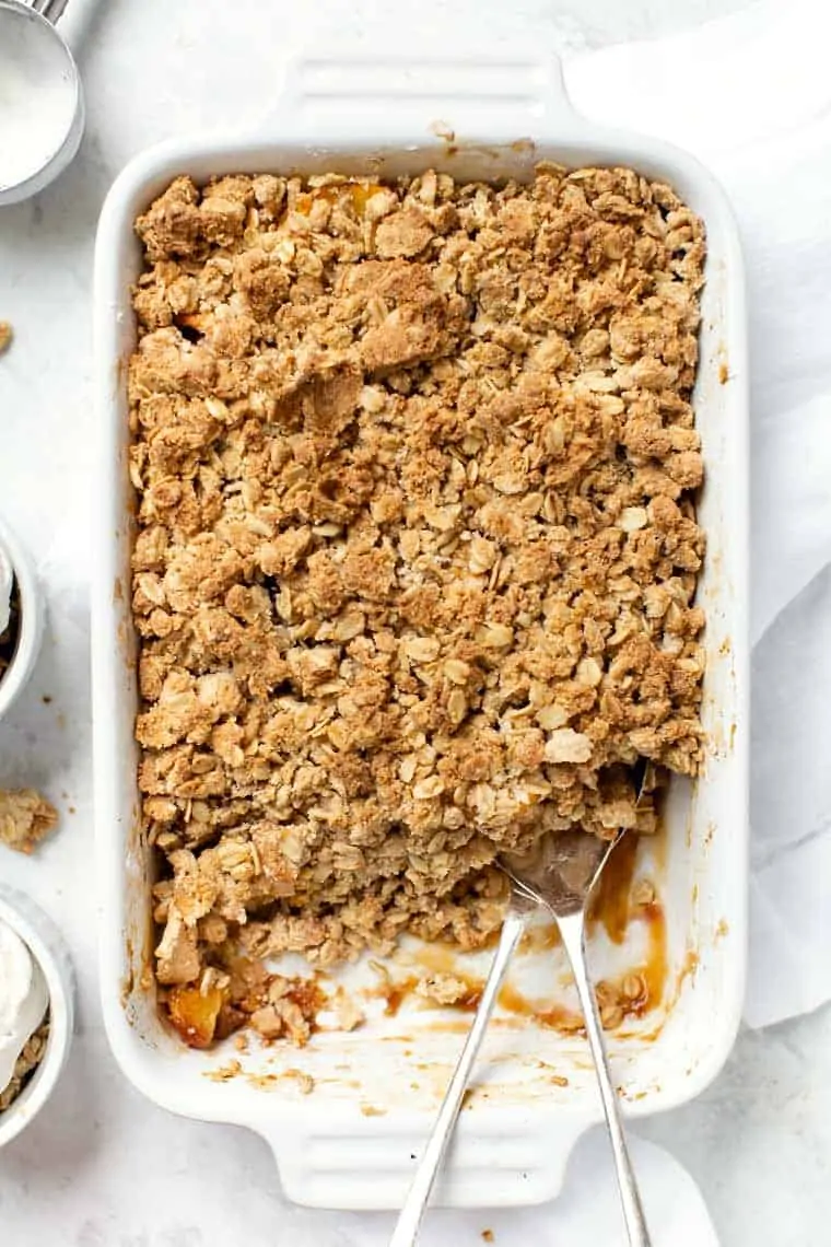 Overhead view of a baking dish with peach crisp, with a few serving taken out and a serving spoon in it