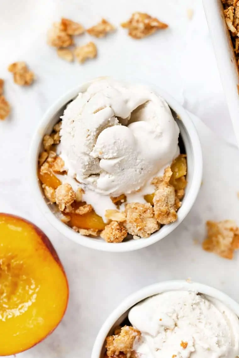 Overhead view of a bowl of peach crisp topped with ice cream, next to another bowl and half a peach