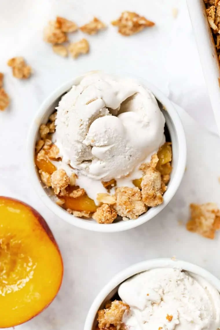 This healthy peach crisp recipe is rich, decadent, perfect for summer, vegan, gluten-free, and only takes 40 minutes to make. Overhead view of a bowl of peach crisp topped with ice cream, next to another bowl and half a peach