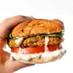 A hand holding up a butternut squash white bean burger in a bun garnished with lettuce, tomato, spicy mayo, and vegan queso.