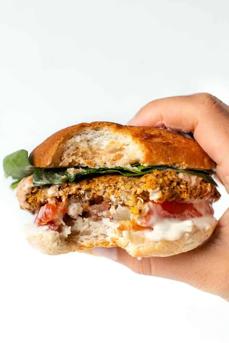 A hand holding up a butternut squash white bean burger in a bun garnished with lettuce, tomato, spicy mayo, and vegan queso, with a bite missing.