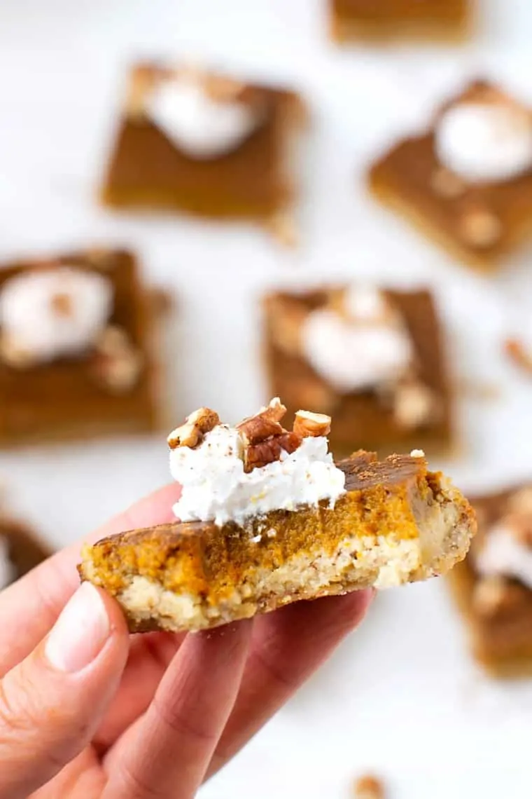 Gluten-, dairy-, and refined sugar-free, these pumpkin pie bars feature a creamy pumpkin pie filling atop a buttery almond flour crust. A hand holding a gluten-free, dairy-free pumpkin pie bar up to the camera.