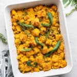 A casserole dish with vegan cornbread stuffing in it, topped with safe, rosemary, and thyme, and fresh herbs around the dish