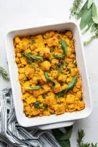 A casserole dish with vegan cornbread stuffing in it, topped with safe, rosemary, and thyme, and fresh herbs around the dish