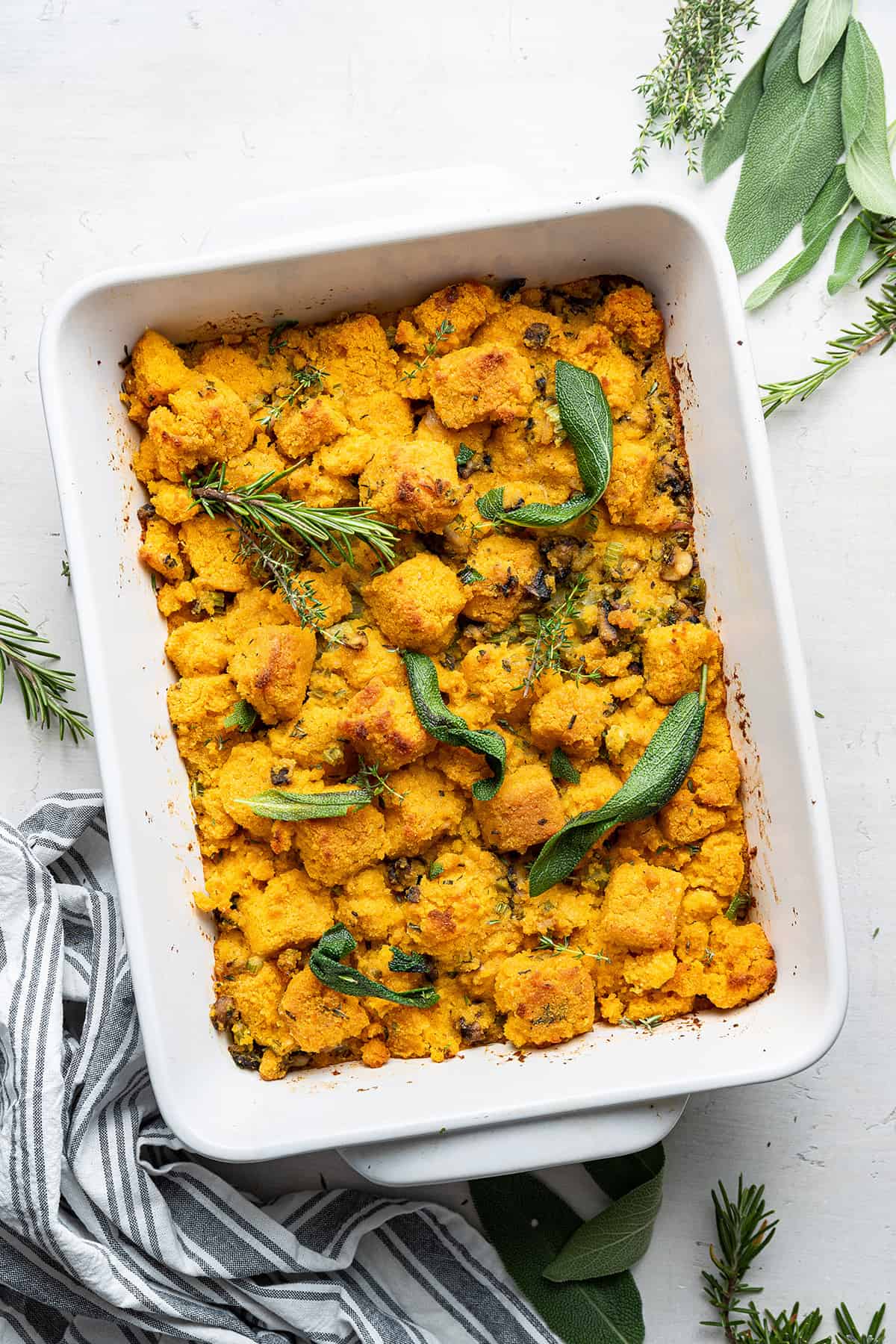This vegan cornbread stuffing recipe is moist and mouthwatering, uses homemade pumpkin cornbread, and is a perfect Thanksgiving side dish A casserole dish with vegan cornbread stuffing in it, topped with safe, rosemary, and thyme, and fresh herbs around the dish
