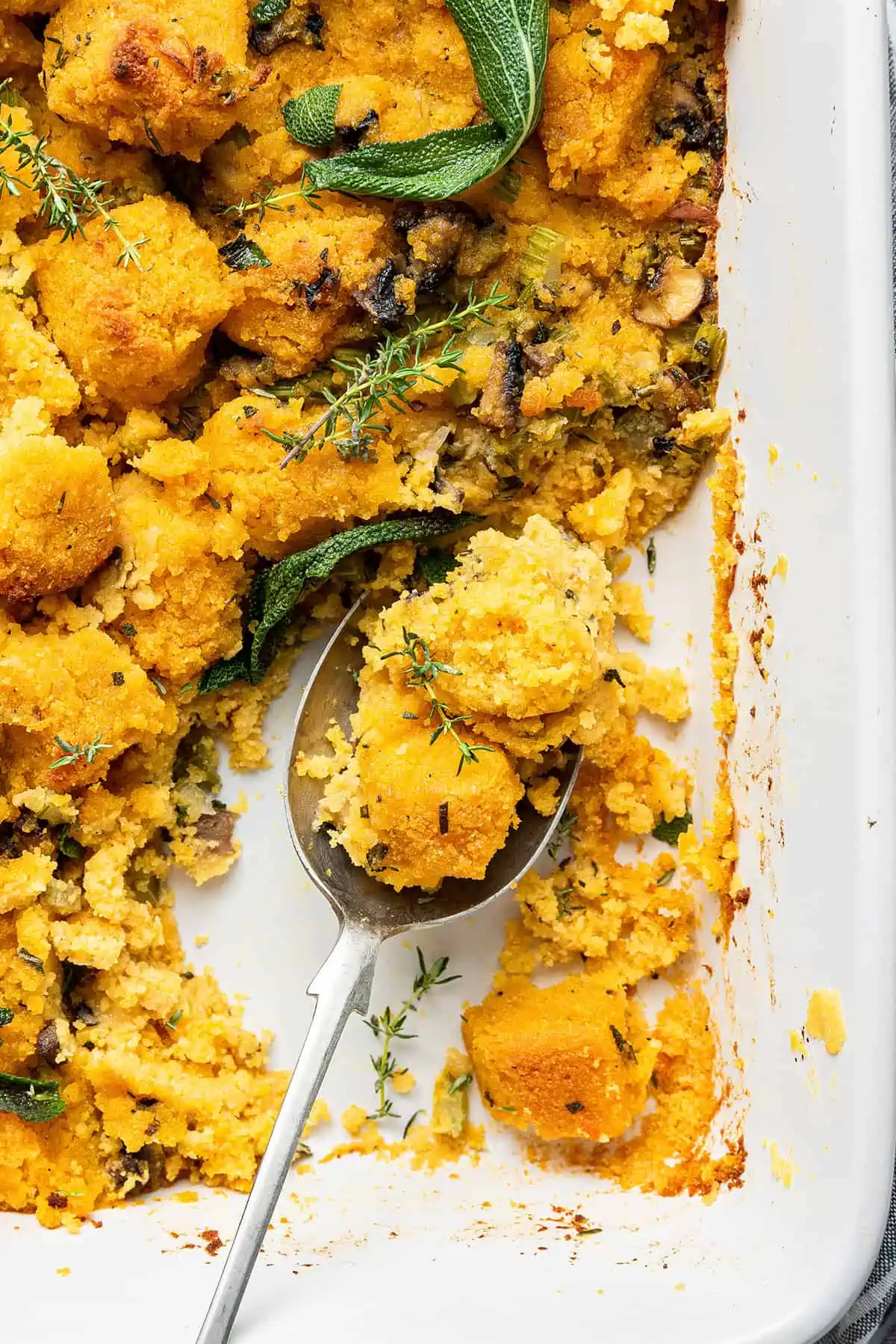 Close up of a serving spoon taking a spoonful of vegan cornbread stuffing, with a thyme sprig on top, out of a casserole dish