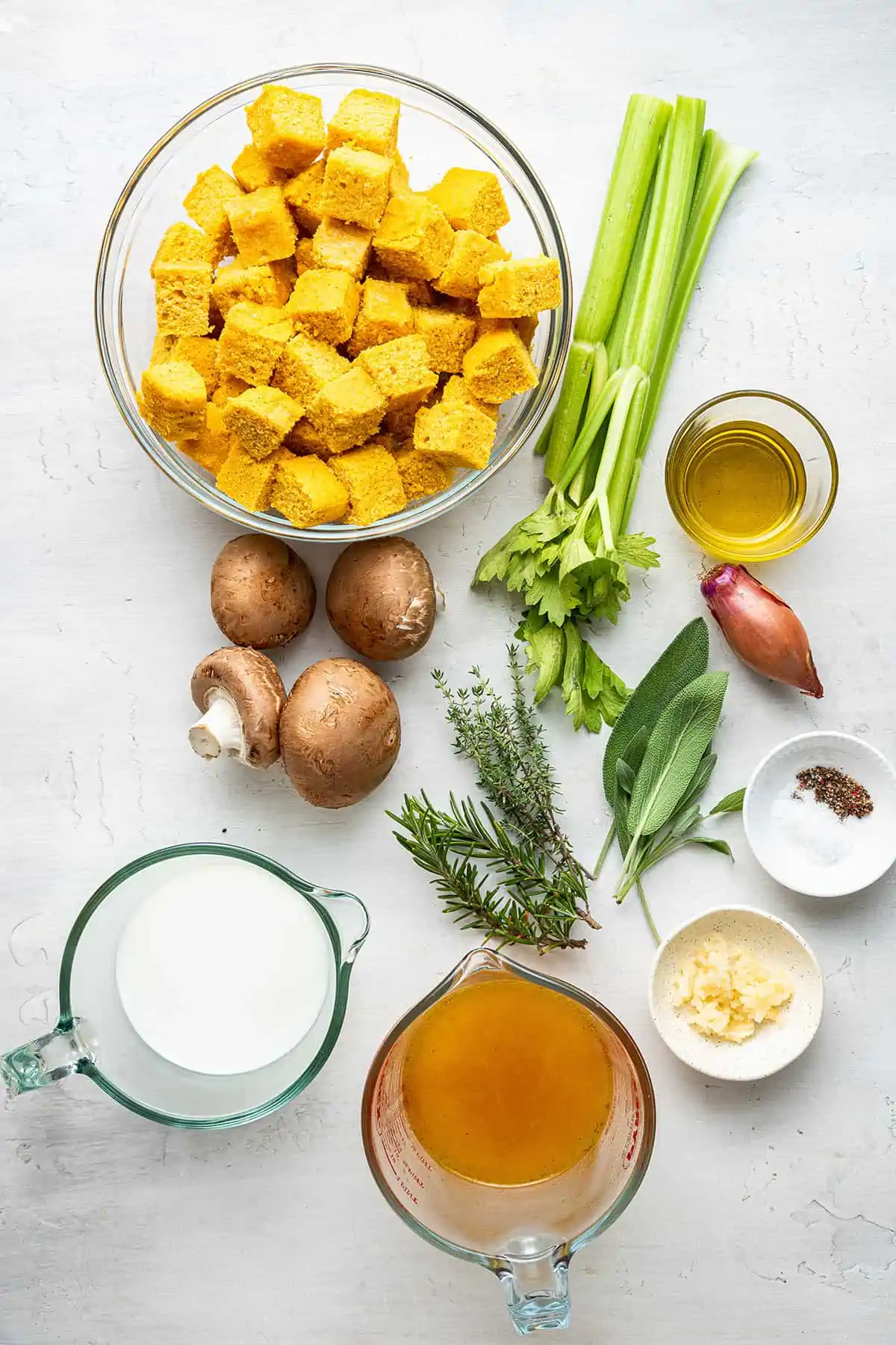 The ingredients for vegan cornbread stuffing: a bowl of cornbread squares, stalks of celery, whole mushrooms, a whole shallot, a bowl of olive oil, a bowl of vegetable broth, a bowl of almond milk, a bowl of salt and pepper, a bowl of minced garlic, and fresh thyme, rosemary, and sage