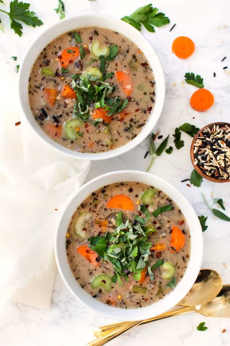 Overhead view of two bowl of wild rice soup, garnished with basil and chili flakes, with slices of carrots and parsley leaves on the table around them, and a bowl of uncooked wild rice