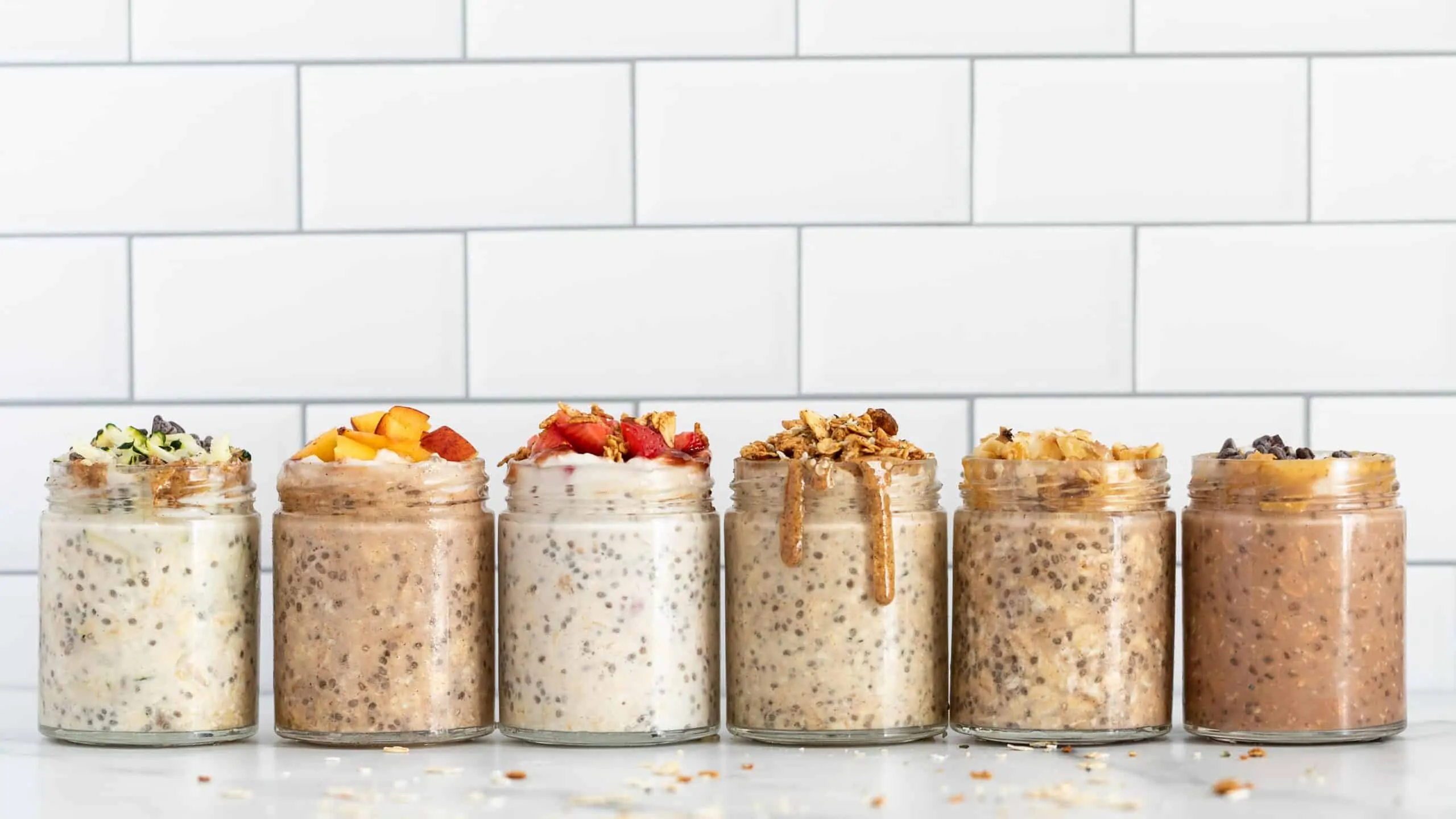 6 mason jars on a marble counter filled with overnight oats and various toppings