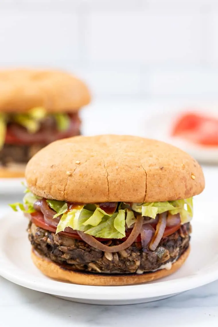 This vegan mushroom veggie burger with white beans is the best you'll ever try! Meaty, easy to make, perfectly seasoned, and gluten-free! A mushroom veggie burger on a plate with a second burger in the background.