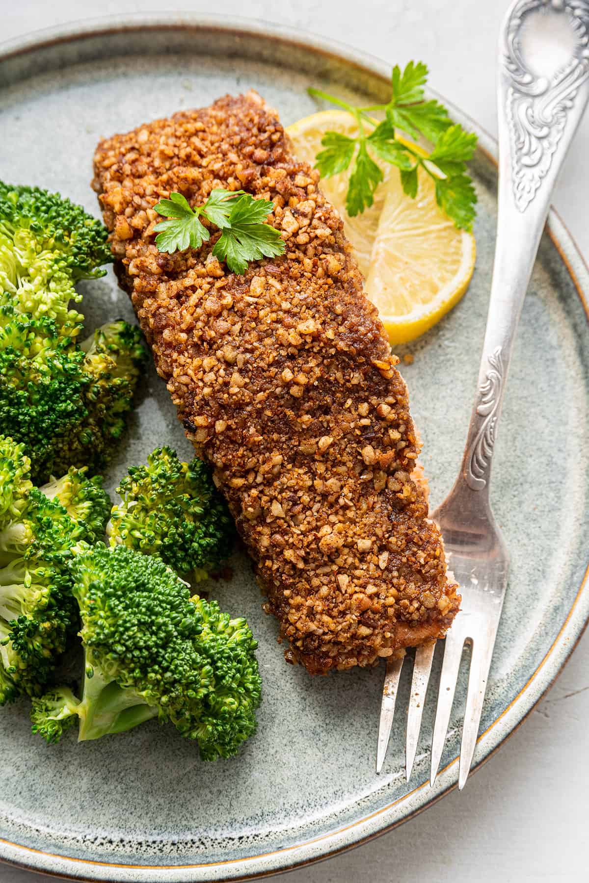 A serving of pecan crusted salmon next to a fork on a plate with a side of broccoli and a lemon wedge.