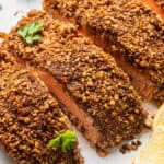 Four pecan crusted salmon fillets on a plate next to a lemon wedge.