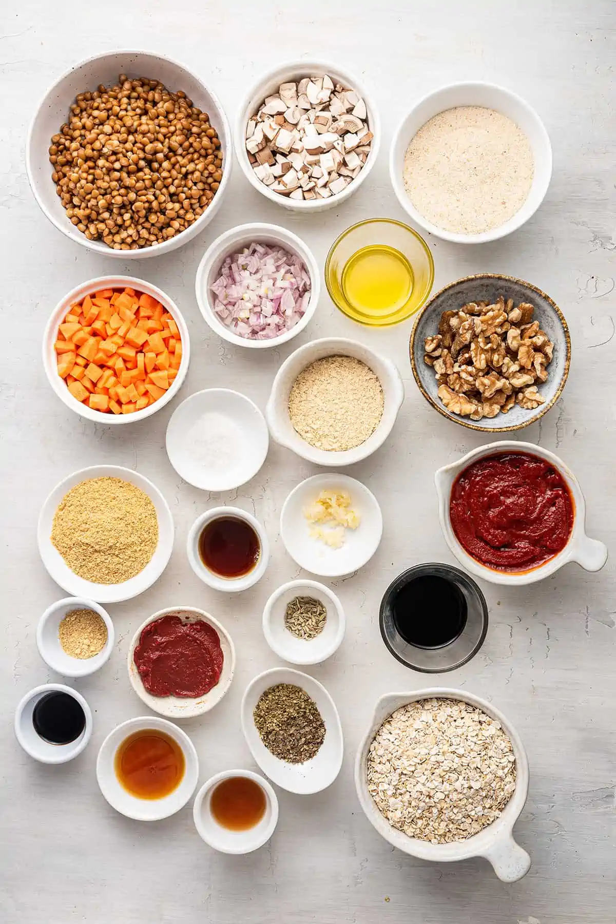 Overhead view of the ingredients for vegan meatloaf, each in a separate bowl: lentils, mushrooms, bread crumbs, carrots, shallots, oil, walnuts, nutritional yeast, tomato paste, vinegar, maple syrup, tamari, ground mustard, oats, garlic, salt, pepper, flaxseed meal, hot sauce, oregano, basil, and fennel seeds