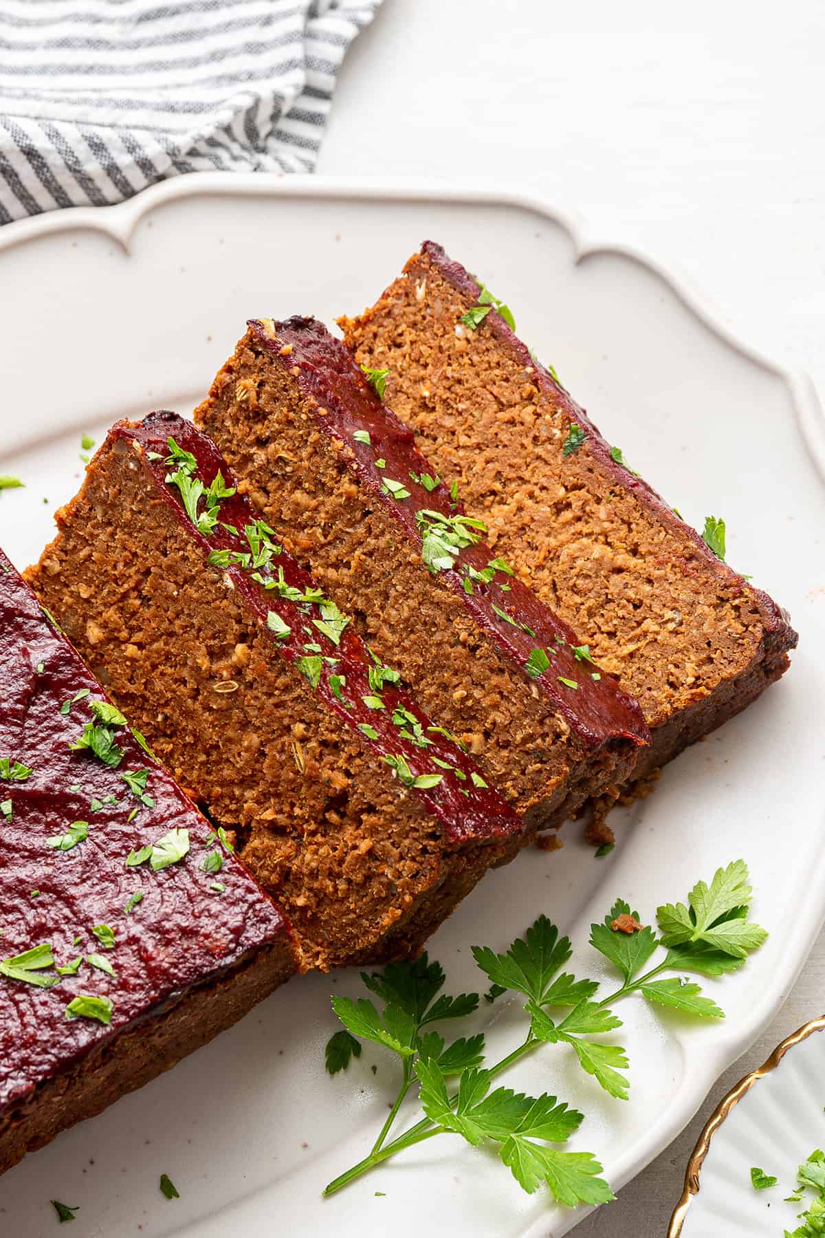 This gluten-free, vegan meatloaf is moist, hearty, comforting, and packed with lentils. It's topped with a sticky, sweet glaze. Close up of three slices of vegan meatloaf on a serving tray, next to the rest of the meatloaf and some parsley