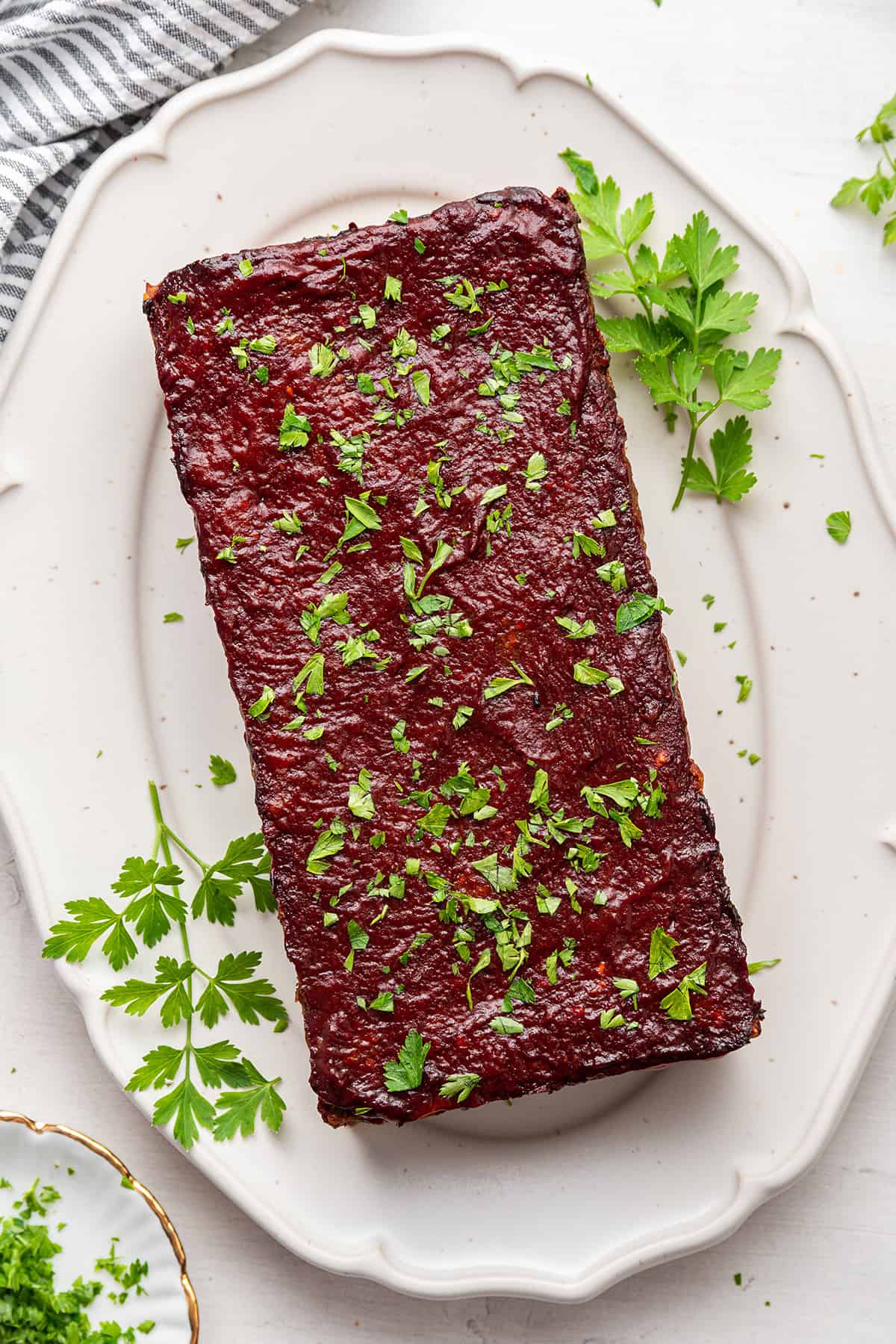 This gluten-free, vegan meatloaf is moist, hearty, comforting, and packed with lentils. It's topped with a sticky, sweet glaze. A vegan meatloaf on a serving tray topped with chopped parsley next to sprigs of parsley