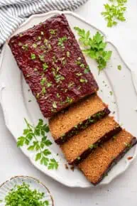 A serving tray with half a vegan meatloaf on it, uncut, and three slices of vegan meatloaf, topped with parsley, with parsley on the serving tray