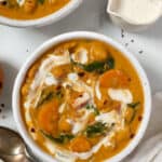 Overhead view of a bowl of chickpea and pumpkin stew swirled with vegan cream, next to a spoon, a creamer jug, and a second bowl of stew.