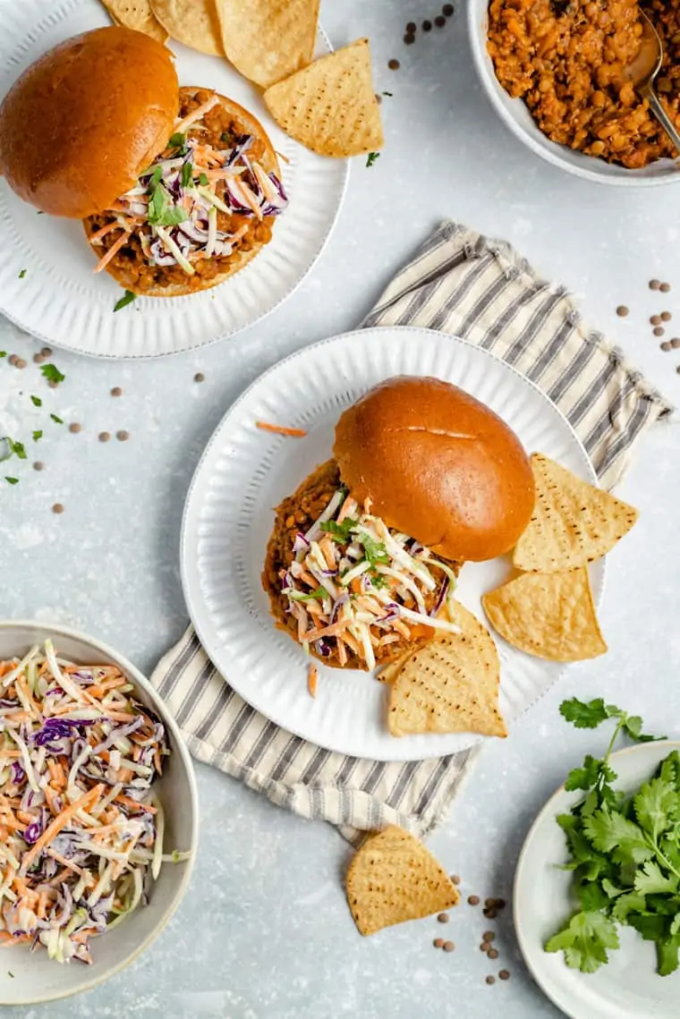 Overhead view of two plates with lentil sloppy joe sandwiches with the top bun to the side, slaw on the sandwiches, and tortilla chips on the side, next to a bowl of sloppy joes mixture, a bowl of slaw, and a plate of cilantro