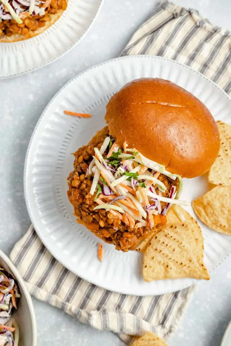 Overhead view of a lentil sloppy joes sandwich on a plate, with the top bun to the side, slaw on the sandwich, and tortilla chips on the plate