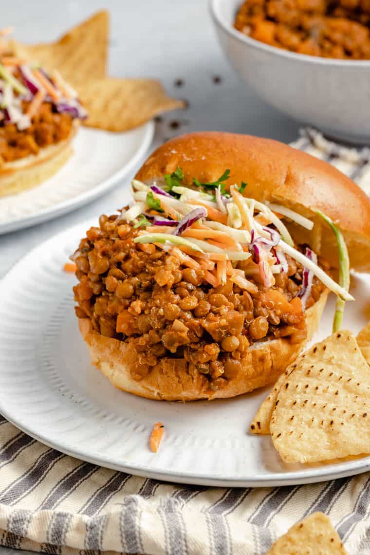 Close up on vegan lentil sloppy joe filling on a hamburger bun with tortilla chips and carrot slaw on a white plate with a stripped dish towel
