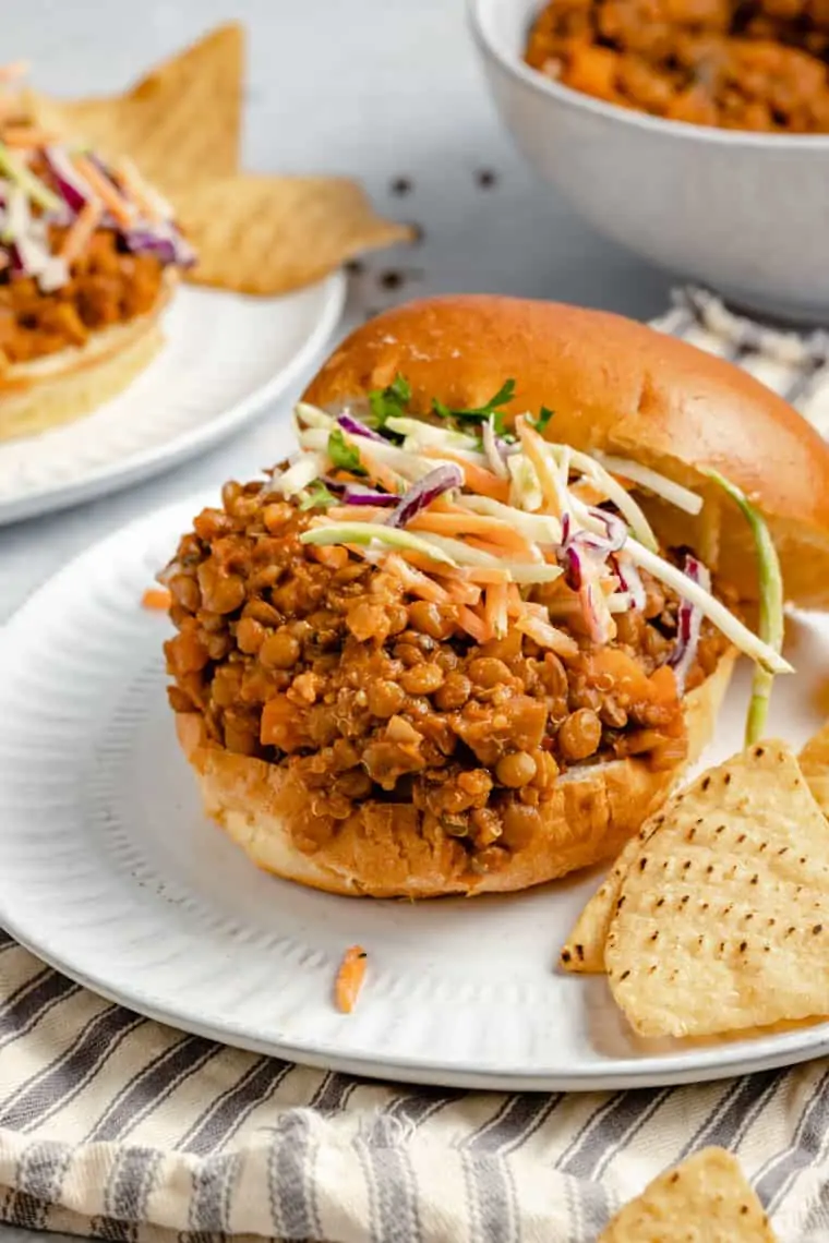 A plate with a lentil sloppy joes sandwich with the bun to the side, slaw on the sloppy joes, and tortilla chips on the plate