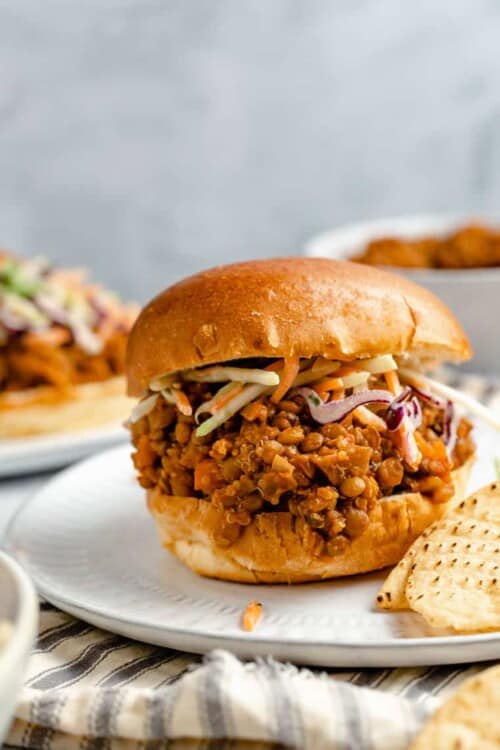 Vegan lentil sloppy joe sandwich on a hamburger bun with tortilla chips and vegetable slaw on a white plate with a stripped dishcloth