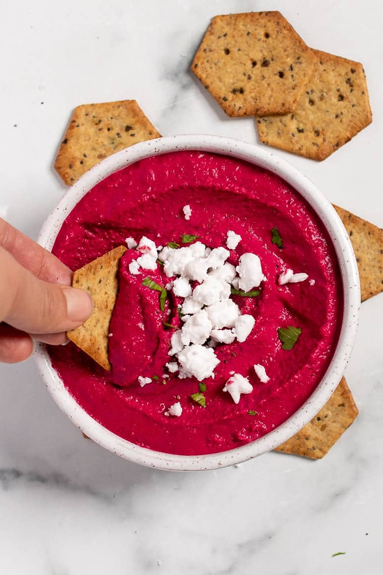 Learn how to make hummus at home, plus easy recipe variations packed with flavorful ingredients like roasted red peppers, beets, and pesto! Overhead view of a hand dipping a cracker into a bowl of beet and feta hummus.