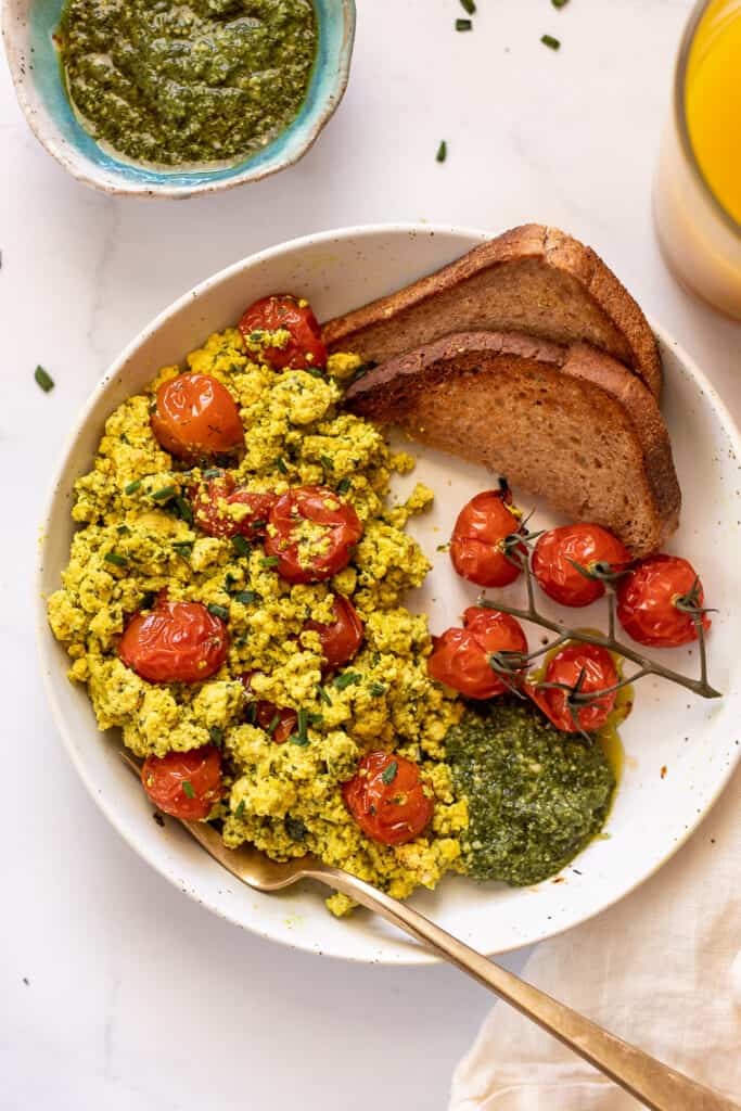 This 10-minute tofu scramble recipe is a satisfying plant-based breakfast with nutritional yeast, turmeric, and garlic for spice and flavor. Overhead view of a pesto tomato tofu scramble with roasted tomatoes and two slices of toast on a plate.