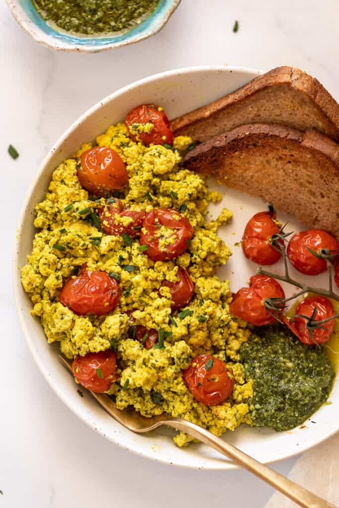 This 10-minute tofu scramble recipe is a satisfying plant-based breakfast with nutritional yeast, turmeric, and garlic for spice and flavor. Overhead view of a pesto tomato tofu scramble with roasted tomatoes and two slices of toast on a plate.