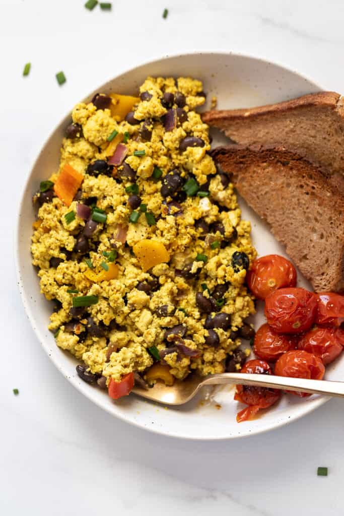 This 10-minute tofu scramble recipe is a satisfying plant-based breakfast with nutritional yeast, turmeric, and garlic for spice and flavor. Southwestern tofu scramble on white plate with tomatoes and toast on the side.