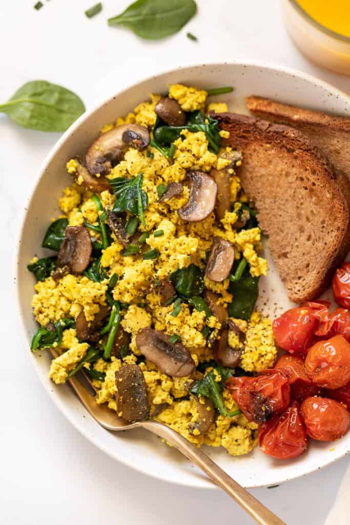 This 10-minute tofu scramble recipe is a satisfying plant-based breakfast with nutritional yeast, turmeric, and garlic for spice and flavor. Overhead view of mushroom and spinach tofu scramble with roasted tomatoes and two slices of toast on a plate.