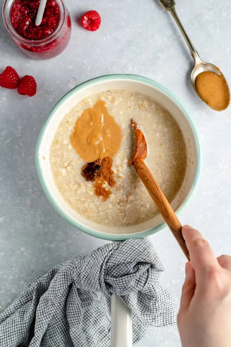 overhead of a pot with oats and peanut butter and dried seasonings stirred in by a hand