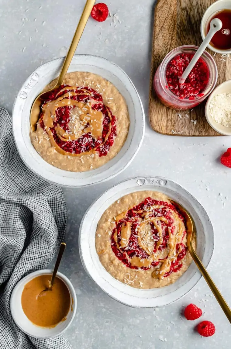 overhead of two white bowls with oatmeal topped with peanut butter and jelly with fresh raspberries on the side