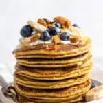 Stack of banana protein pancakes with fresh blueberries, maple syrup, almonds and bananas on top