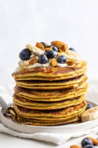Stack of banana protein pancakes with fresh blueberries, maple syrup, almonds and bananas on top