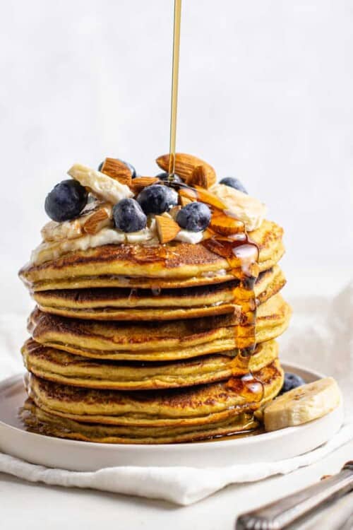 stack of protein pancakes topped with fruit and nuts, being drizzled with maple syrup
