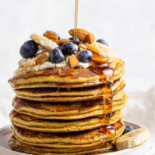 stack of protein pancakes topped with fruit and nuts, being drizzled with maple syrup