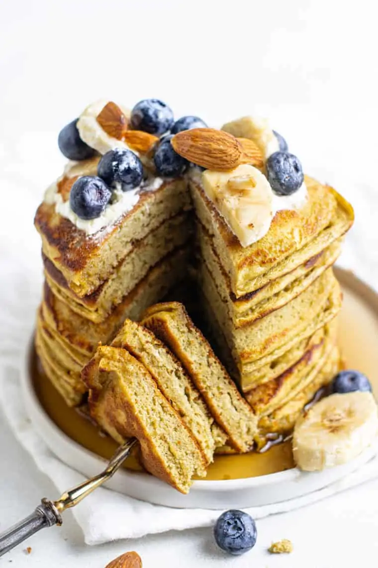 stack of protein pancakes with blueberries, almonds and banana slices on a white plate with a slice removed by a fork