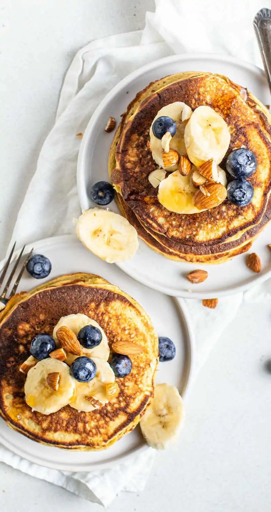 Overhead shot of protein pancakes stacked on two plates with bananas, blueberries, and almonds