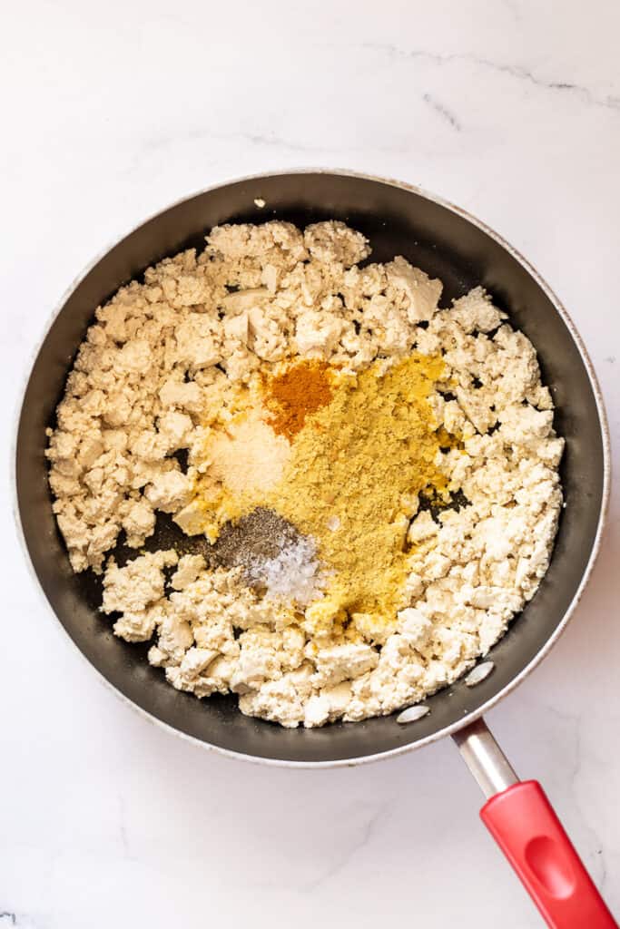 This 10-minute tofu scramble recipe is a satisfying plant-based breakfast with nutritional yeast, turmeric, and garlic for spice and flavor. Overhead view of spices added to crumbled tofu in a skillet.