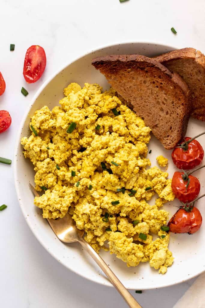 This 10-minute tofu scramble recipe is a satisfying plant-based breakfast with nutritional yeast, turmeric, and garlic for spice and flavor. Overhead view of a tofu scramble with roasted tomatoes and two slices of toast on a plate.