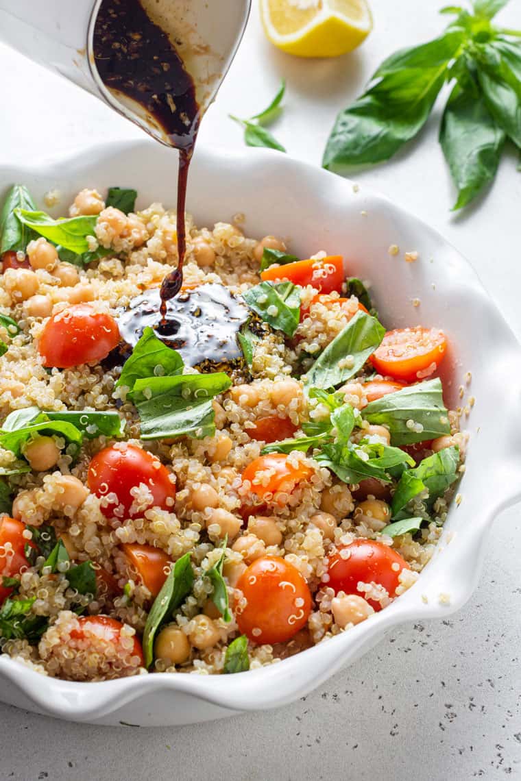 pouring balsamic vinegar dressing on quinoa salad with tomatoes and basil