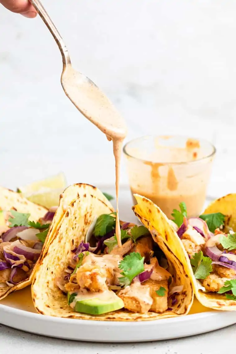 pouring tahini sauce over three crispy fish tacos