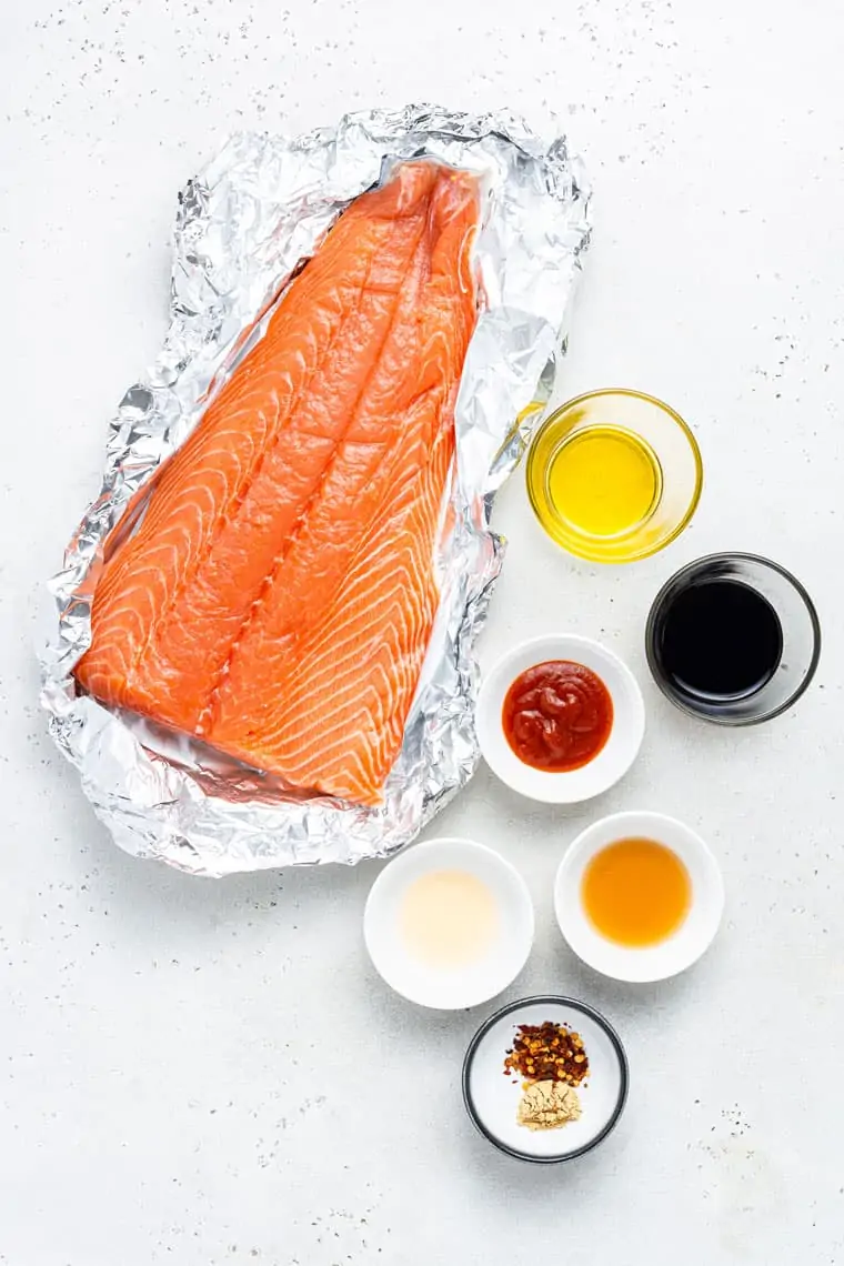 large piece of salmon in a foil wrapper with ingredients for sauce in small bowls on the side