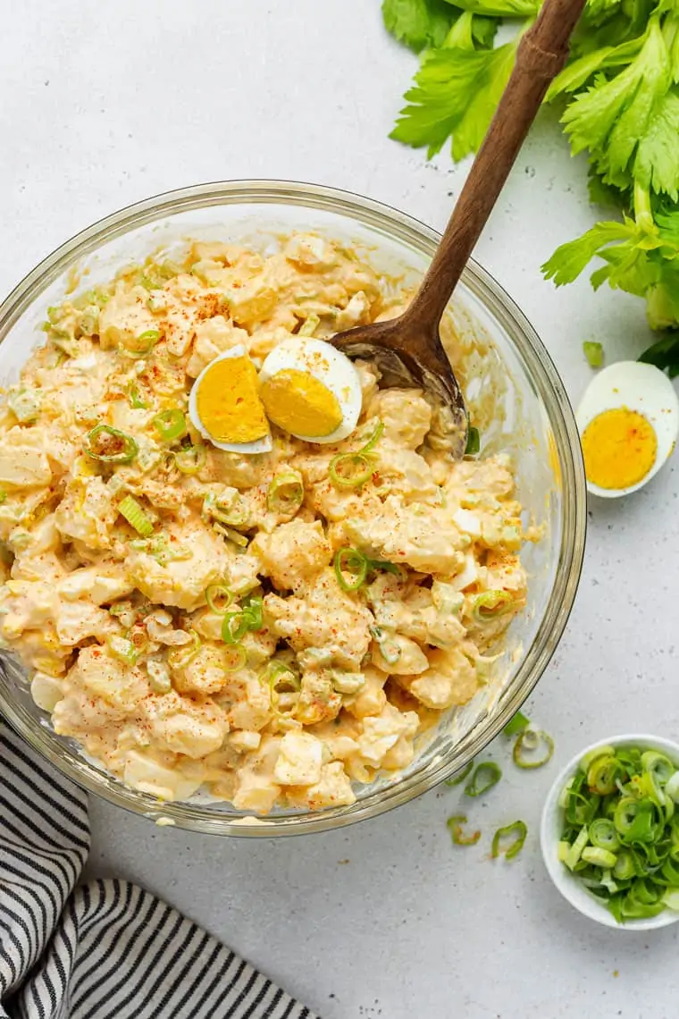 This healthy potato salad recipe is the perfect summer side dish, and goes will with any barbecue, outdoor party, or picnic. Overhead view of a bowl of potato salad topped with scallions, paprika, and a boiled egg, with a wooden spoon in it, next to a bowl of scallions, half a boiled egg, and some celery
