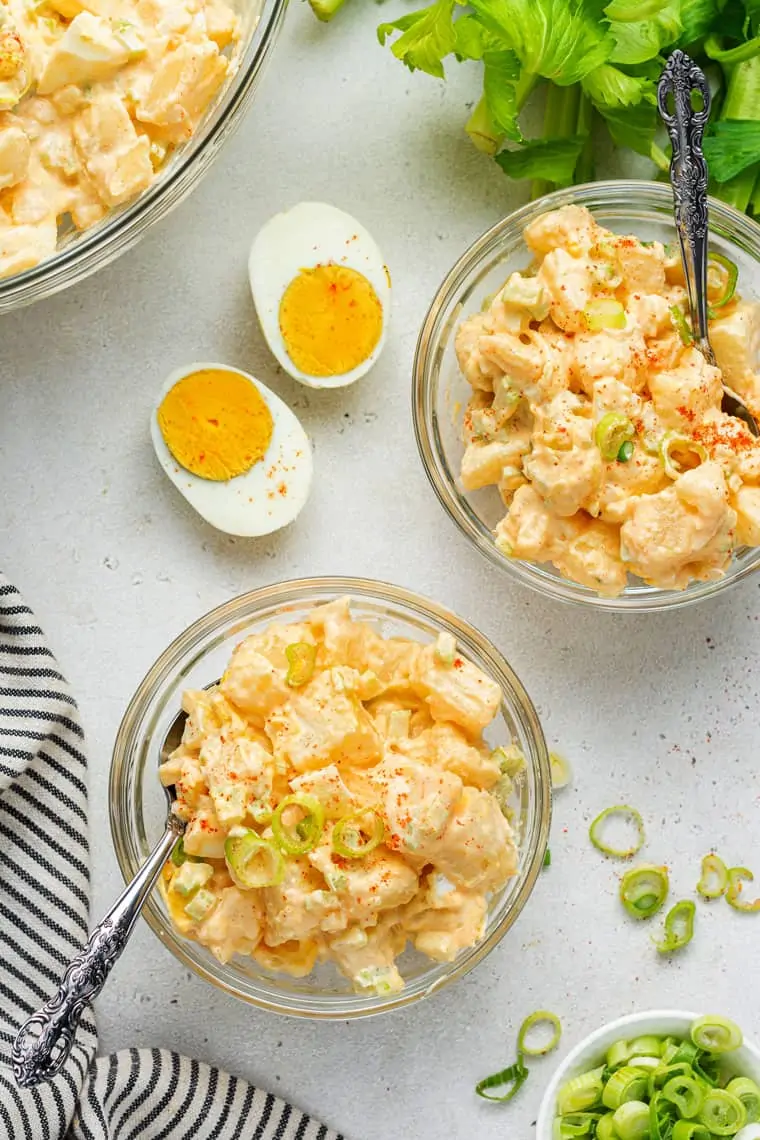 Overhead view of two bowls of potato salad topped with scallions, with spoons in them, next to a hard boiled egg cut in half