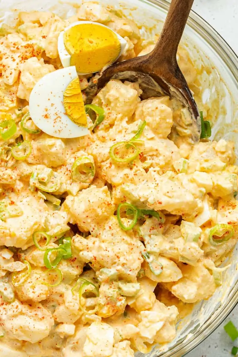 Close up of potato salad topped with scallions, paprika, and a hard boiled egg, with a wooden spoon in it