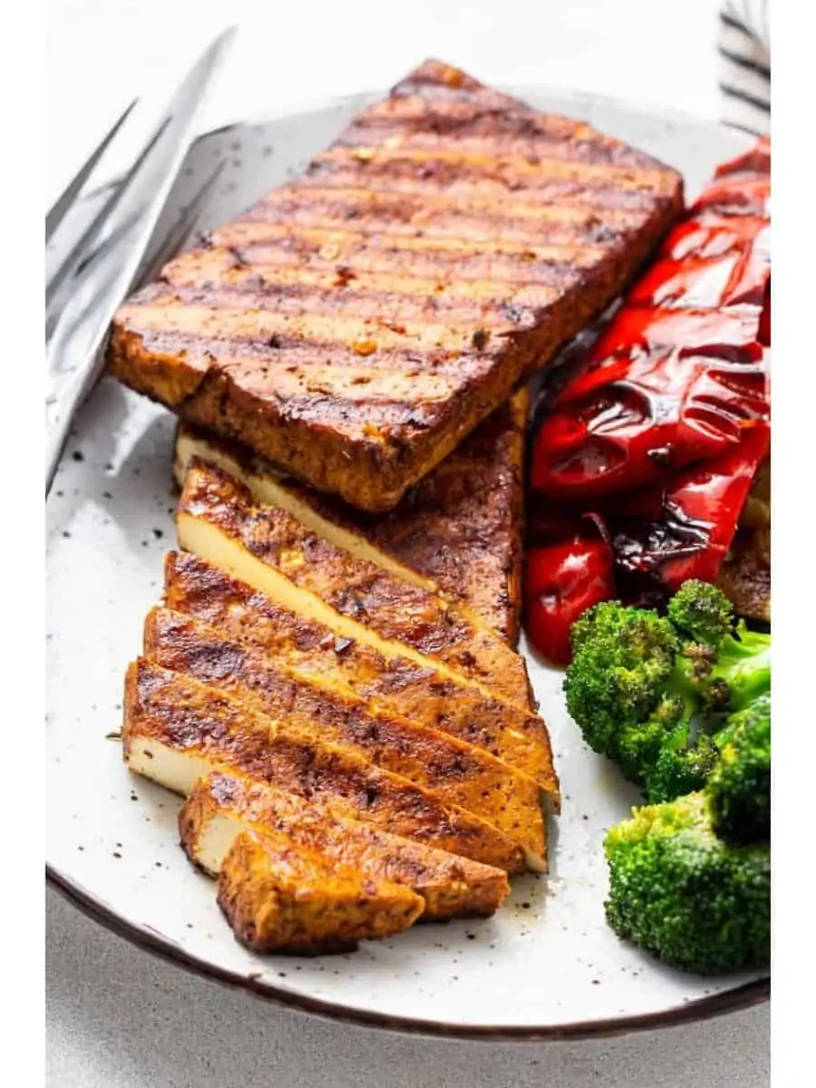 Two slabs of grilled tofu on a plate, with some slices cut, next to roasted peppers and broccoli